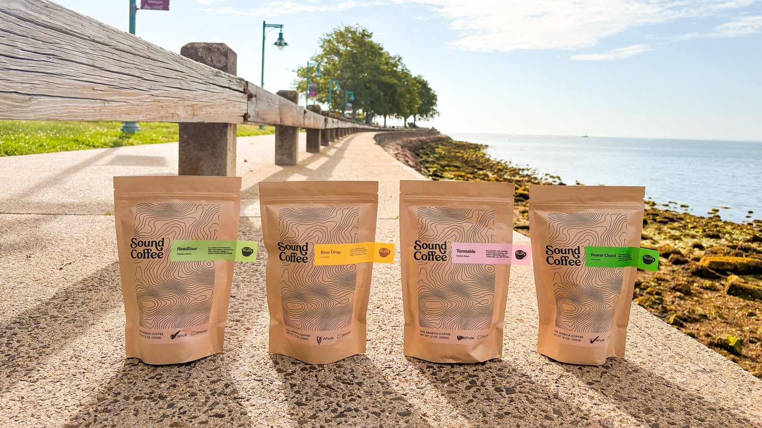 Four stand-up pouches of Sound Coffee in different flavors are lined up on a stone pathway outdoors, with a scenic waterfront walkway, trees, and blue sky in the background.