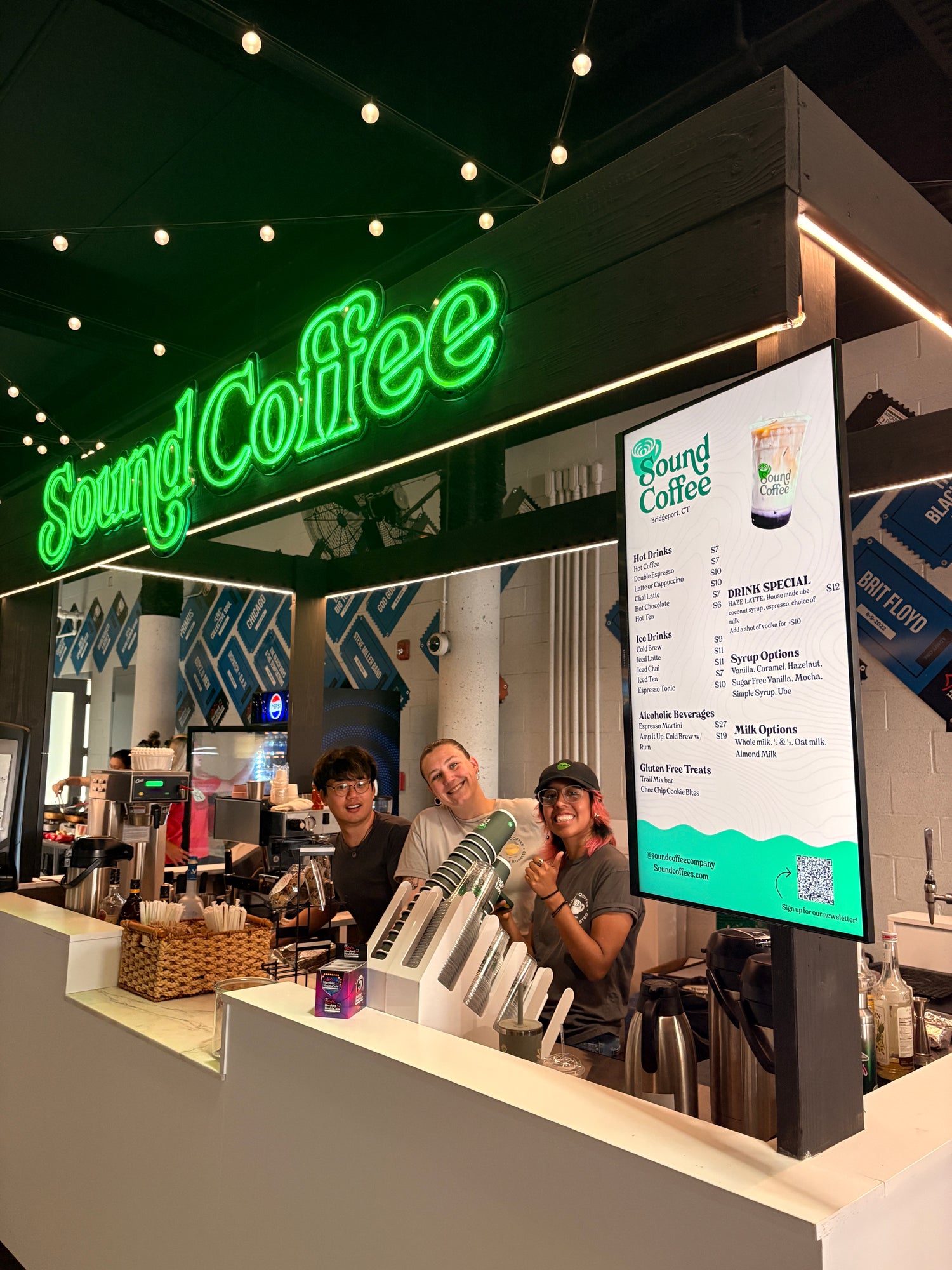 Three baristas stand behind a coffee counter inside a Sound Coffee shop, with a bright green neon sign and a vertical menu board visible above them.